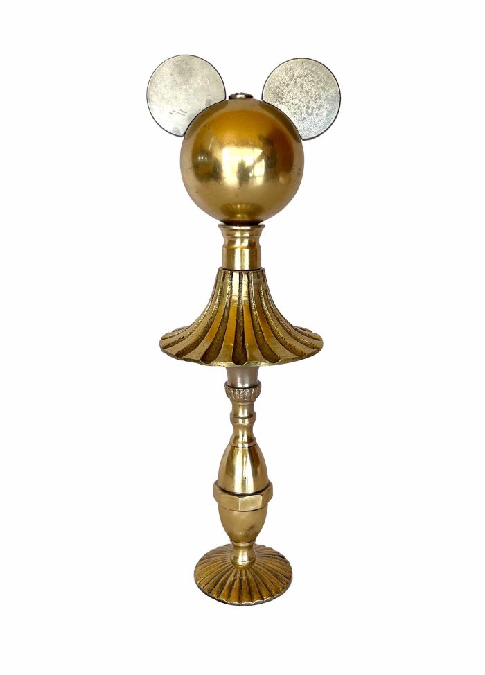Una escultura dorada con un pomo a modo de cara, las orejas hechas con piezas marinas de Cosir y el cuerpo, la falda y las piernas de una lámpara.