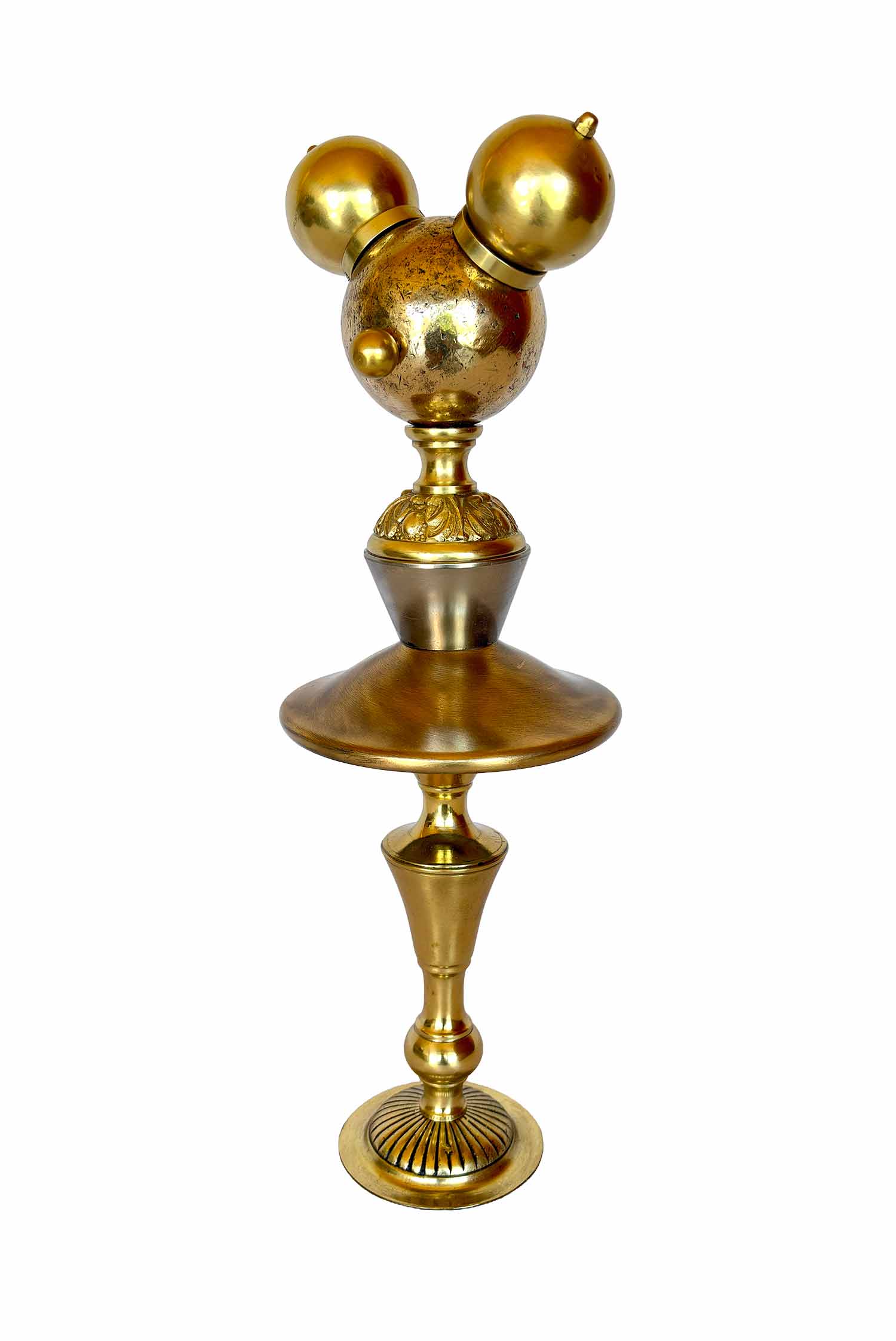 Una escultura dorada con una bola de petanca en cada cara y dos pomos a modo de orejas; el cuerpo y las piernas también están dorados y están hechos con piezas de pantallas de lámpara.
