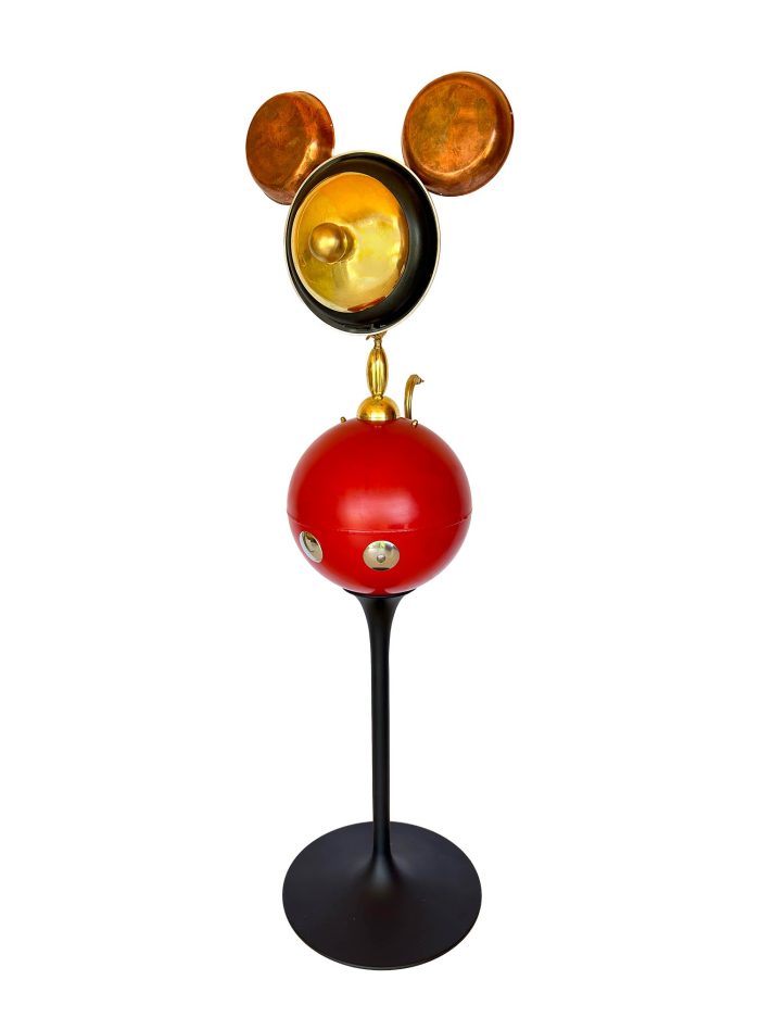 Una escultura en la que el rostro dorado está formado por una sartén y las orejas son una balanza, el cuerpo es una bola roja muy grande y las piernas son una punta tallada en negro.