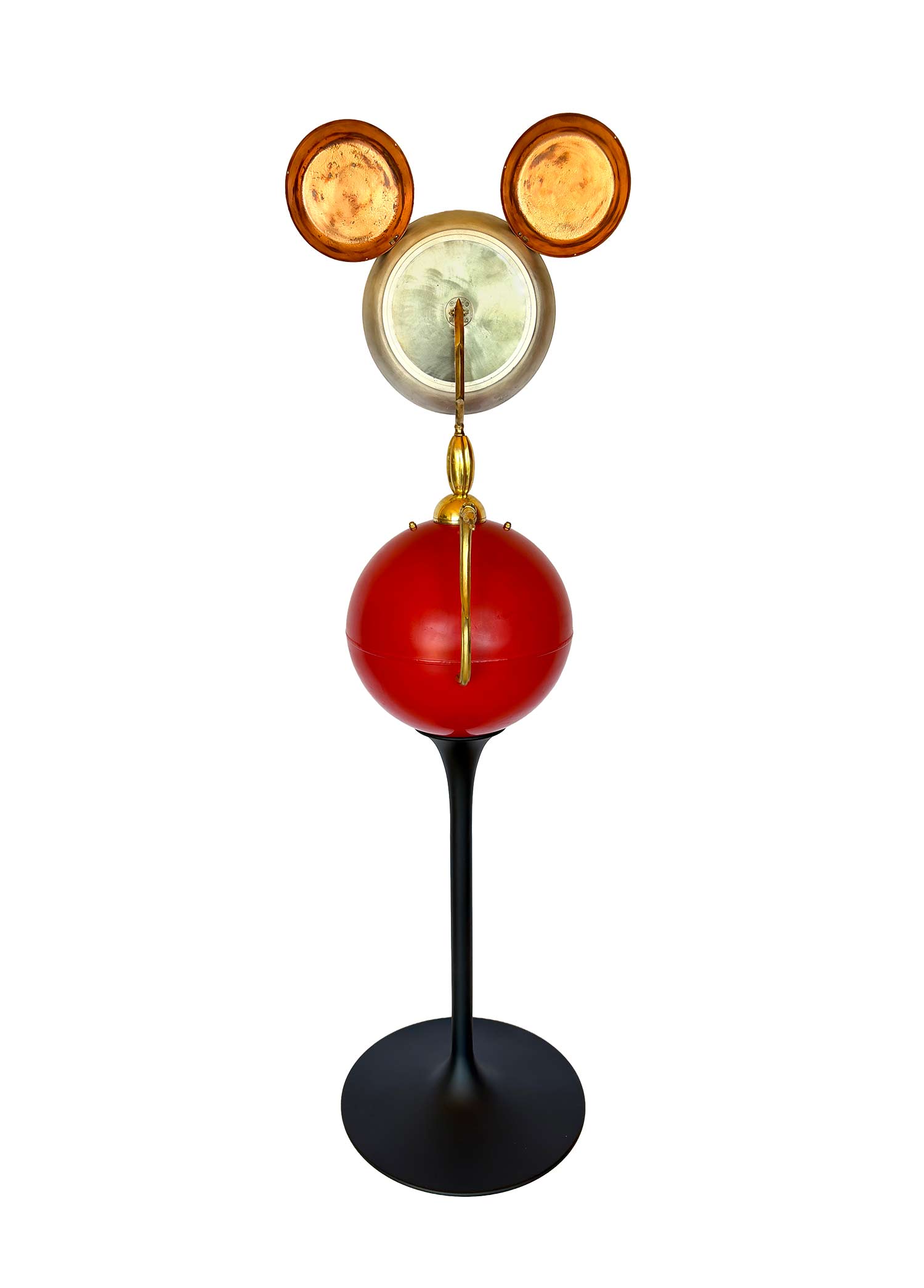 Una escultura en la que el rostro dorado está formado por una sartén y las orejas son una balanza, el cuerpo es una bola roja muy grande y las piernas son una punta tallada en negro.Una escultura en la que el rostro dorado está formado por una sartén y las orejas son una balanza, el cuerpo es una bola roja muy grande y las piernas son una punta tallada en negro.